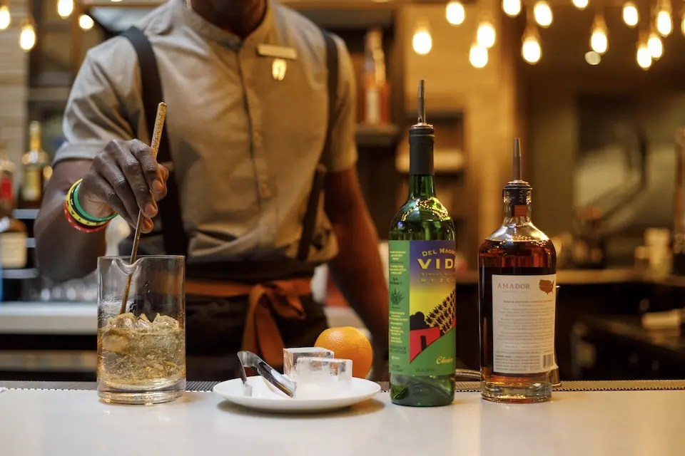 AKB New York - bartender stirring The Redemption cocktail in a mixing glass, with two liquor bottles, ice cubes and an orange on the bar counter