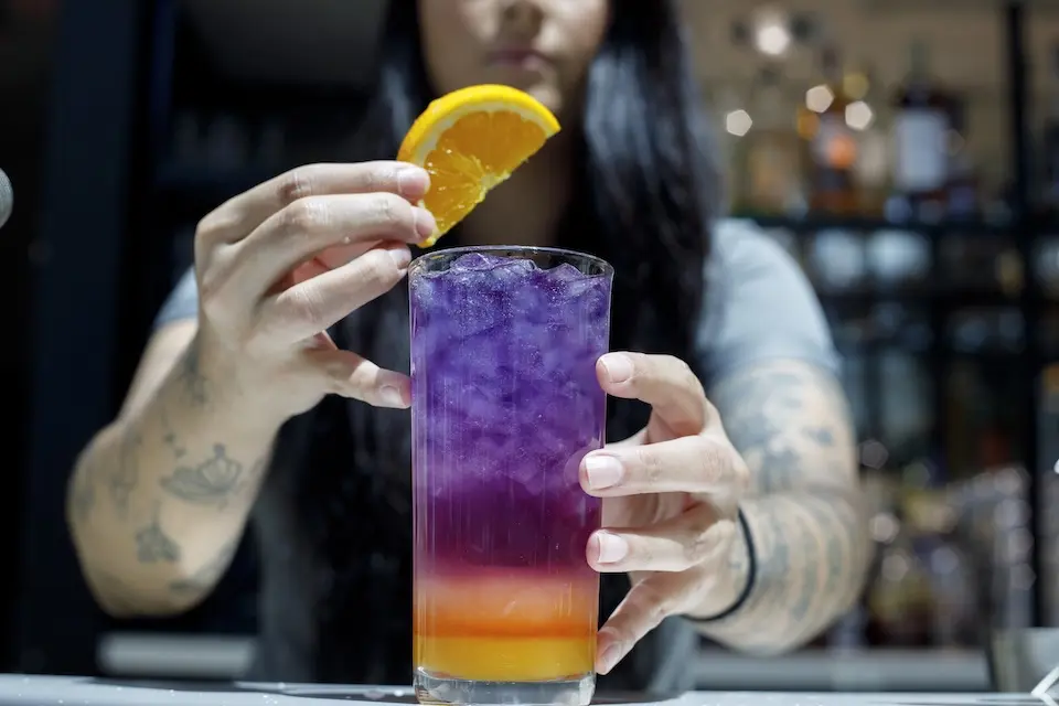 AKB Austin - bartender placing an orange wheel garnish on a Violet Sky cocktail, featuring purple and orange layers over crushed ice