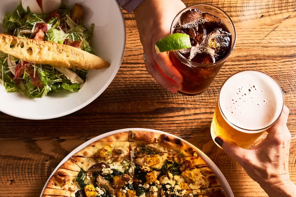 AKB - two diners toasting drinks over flatbread pizza and salad during happy hour