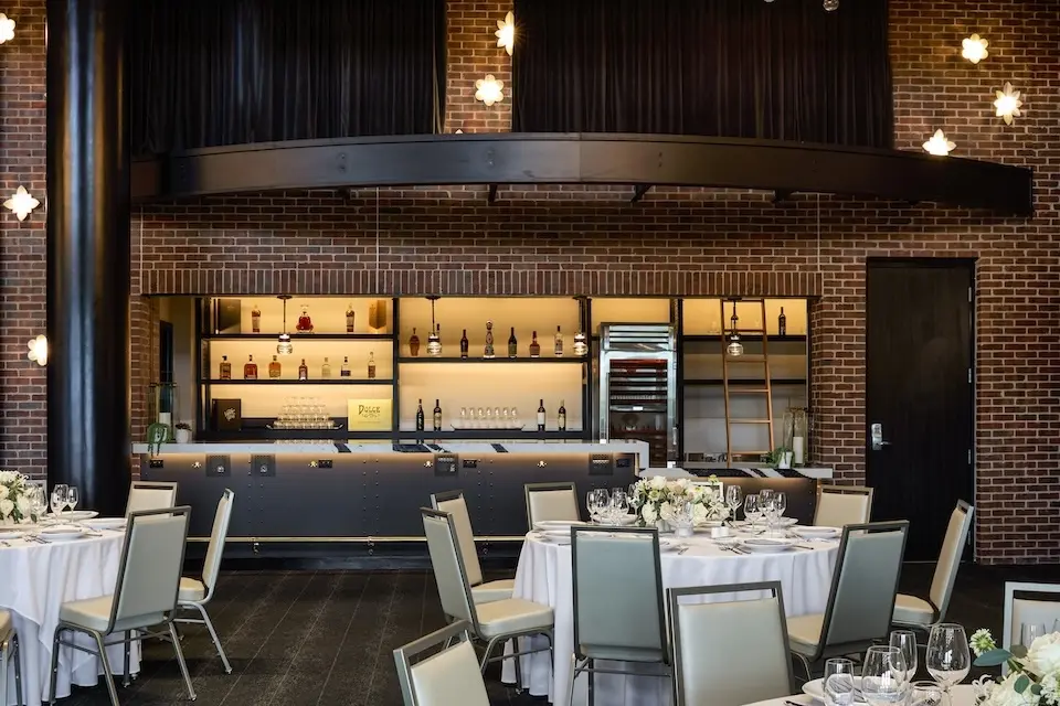 Penthouse - banquet-style event setup with round tables, white linens and high-back chairs in front of a built-in, full-service corner bar