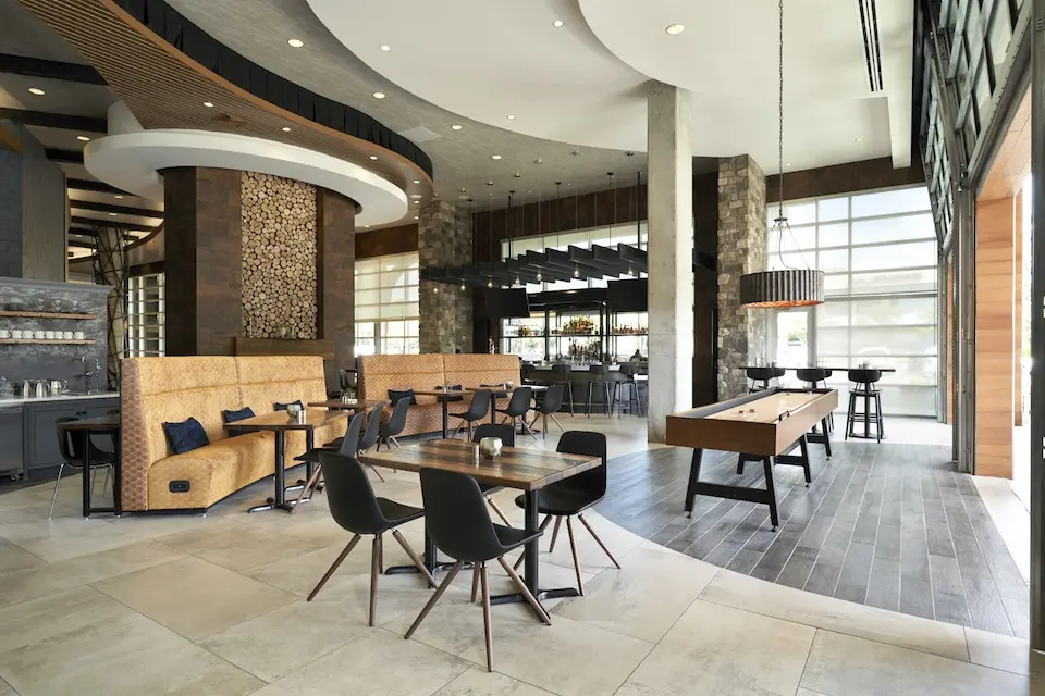 AKB - industrial-chic hotel dining room featuring curved booth seating, wooden dining tables, a shuffleboard table and a cocktail bar