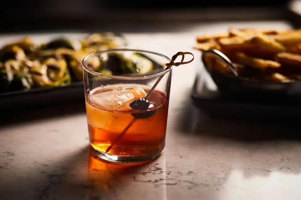 AKB - an old fashioned cocktail with a cherry garnish, set on a bar top with plates of shareable appetizers