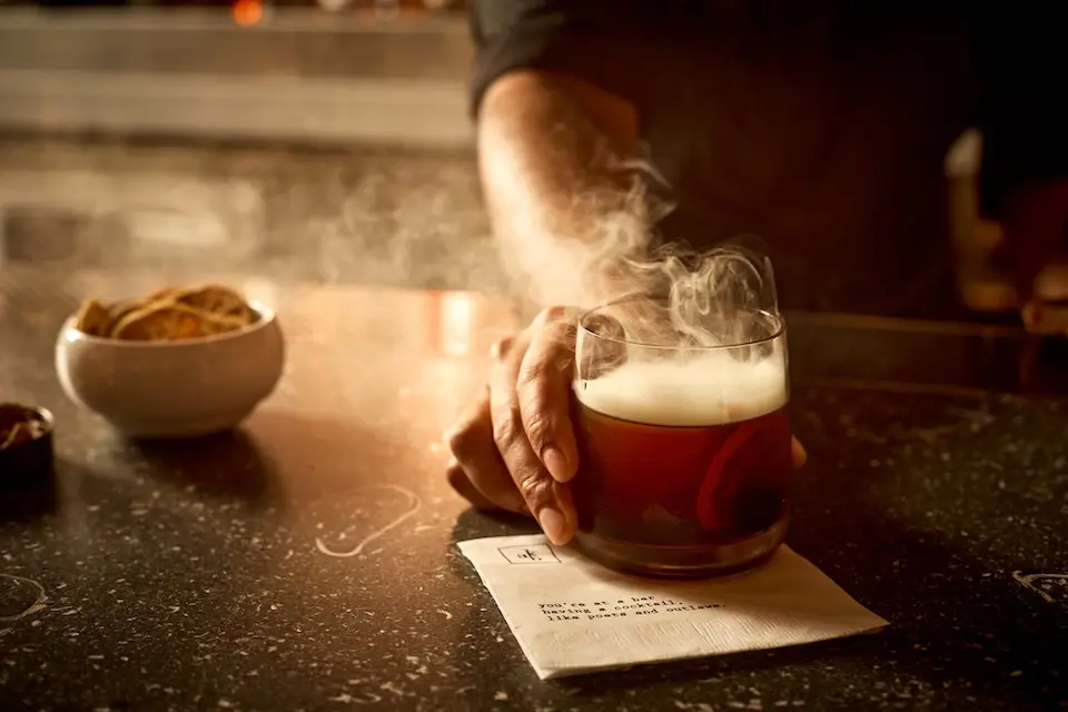 AKB - a bartender serving a smoking Hennessy Smoke Smash cocktail on a napkin over a dark bar counter