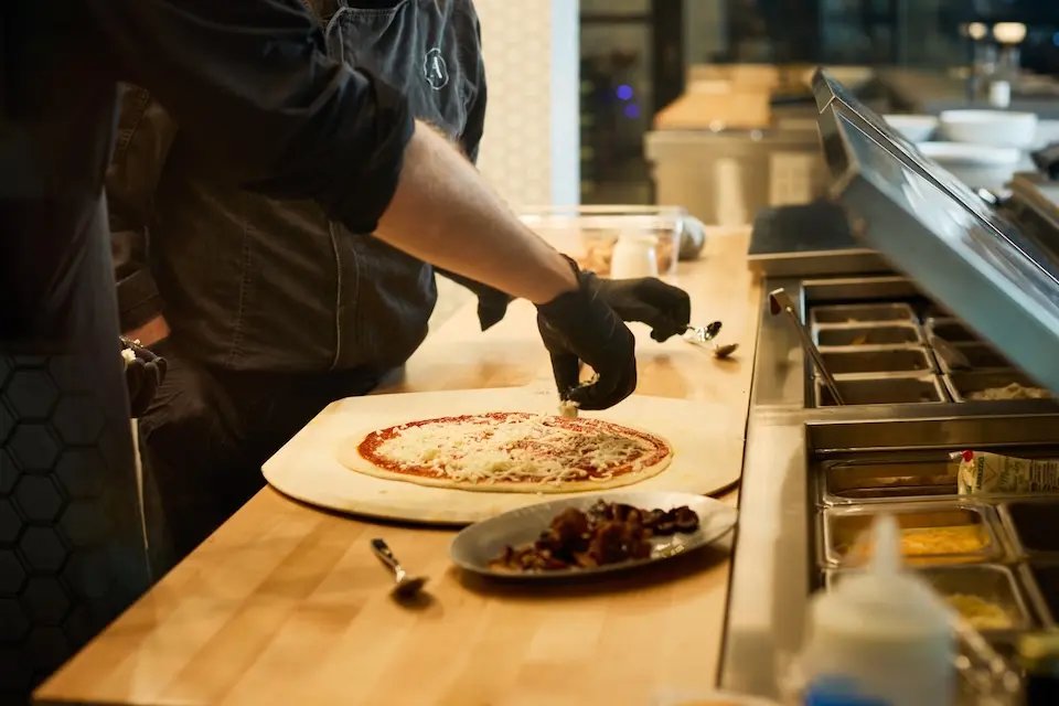 AKB - a chef wearing black gloves adds toppings to a fresh flatbread pizza on a wooden counter in an open kitchen area