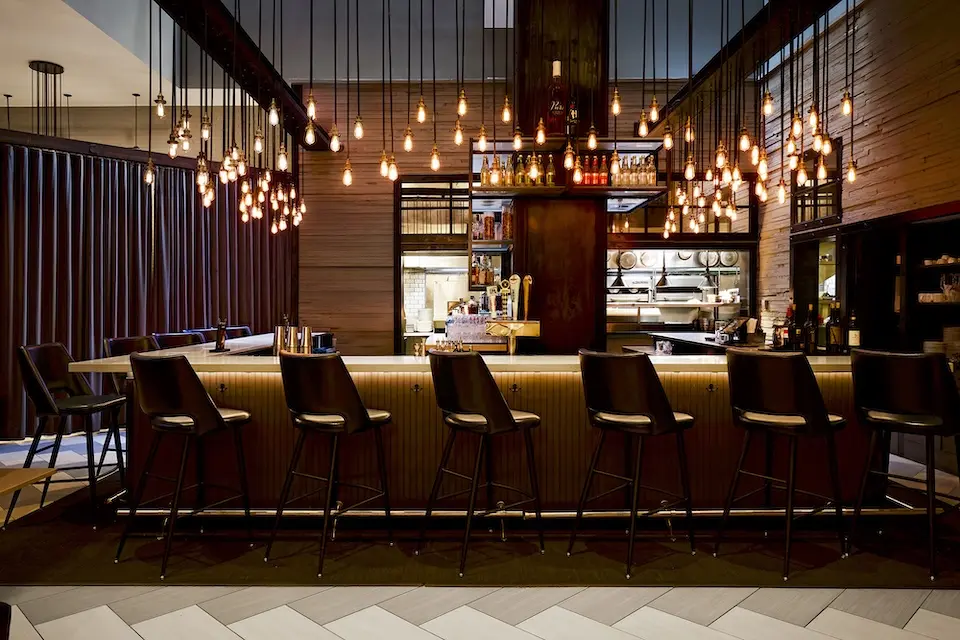 AKB - modern bar counter lined with high-back stools under a dramatic canopy of hanging Edison bulb lights
