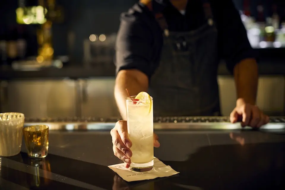 AKB - a bartender serving a tall, clear cocktail garnished with a lemon wheel on a dimly lit bar counter