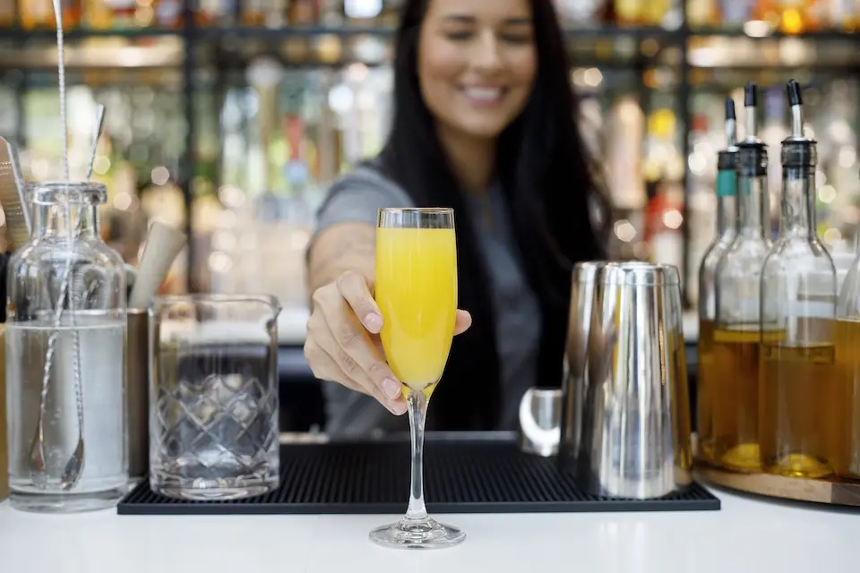 AKB - a female bartender serving a yellow mimosa in a champagne flute on a white bar counter