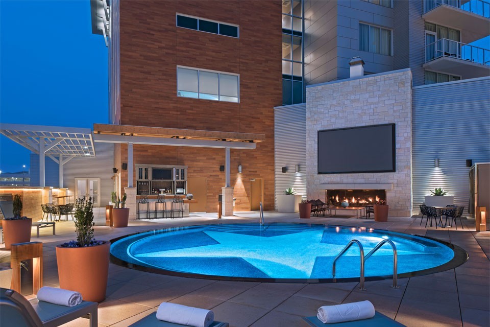 The pool patio at night, with a star-bottom circular pool, giant TV above a fireplace and an outdoor bar