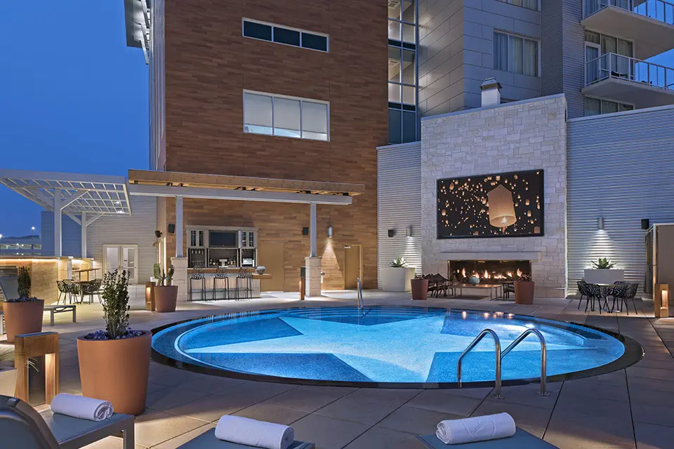 The pool patio at night, with a star-bottom circular pool, giant TV above a fireplace and an outdoor bar